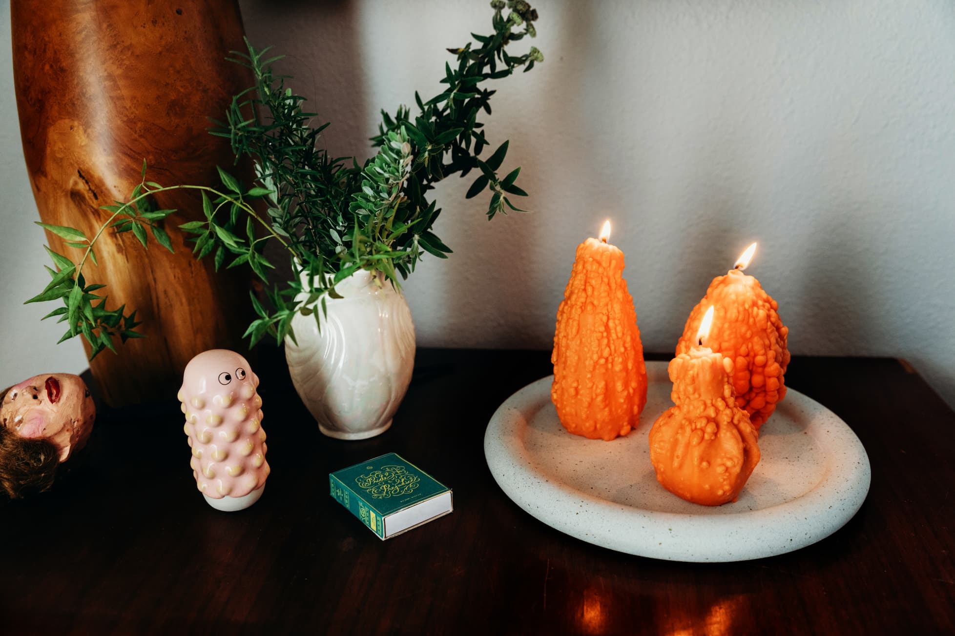 A tablescape with orange gourd-shaped Scandles candles on a white terrazzo tray, a pink bubble matchstick holder, and greenery in a white vase