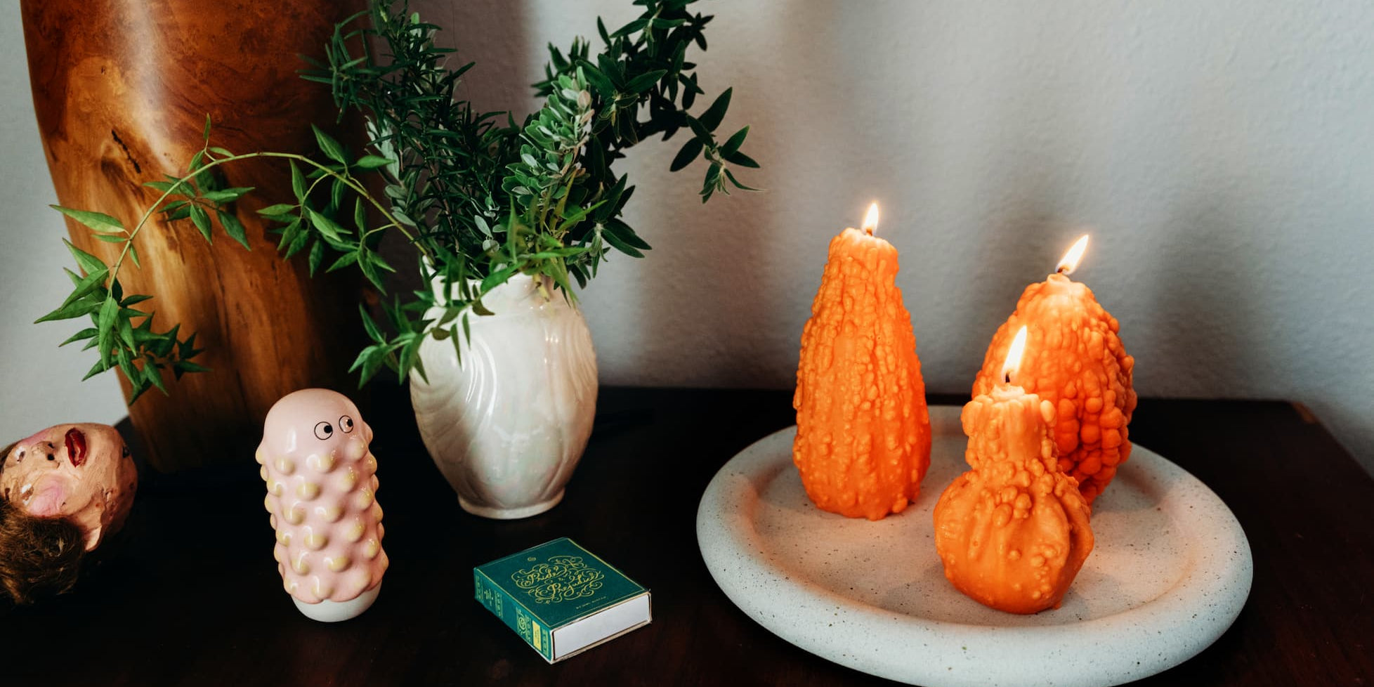A tablescape with orange gourd-shaped Scandles candles on a white terrazzo tray, a pink bubble matchstick holder, and greenery in a white vase